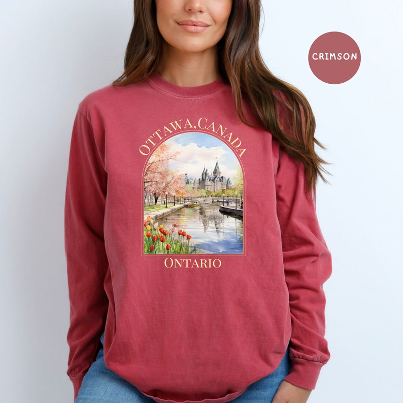 May include: A crimson long-sleeve t-shirt with a graphic print of the Parliament Building in Ottawa, Canada. The graphic features a watercolor style illustration of the building with cherry blossoms and a river in the foreground. The text "OTTAWA, CANADA" is printed in a cream color above the graphic, and the text "ONTARIO" is printed in a cream color below the graphic.