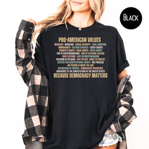 Pode incluir: Camiseta preta com o texto "PRO-AMERICAN VALUES" em dourado e v&aacute;rias cores. O texto lista valores como "Medicare", "LGBTQ+ Rights" e "Liberdade de religi&atilde;o". A frase "BECAUSE DEMOCRACY MATTERS" est&aacute; na parte inferior.
