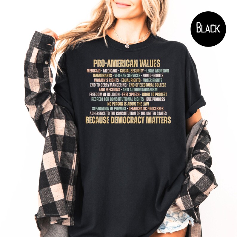 Pode incluir: Camiseta preta com o texto "PRO-AMERICAN VALUES" em dourado e v&aacute;rias cores. O texto lista valores como "Medicare", "LGBTQ+ Rights" e "Liberdade de religi&atilde;o". A frase "BECAUSE DEMOCRACY MATTERS" est&aacute; na parte inferior.