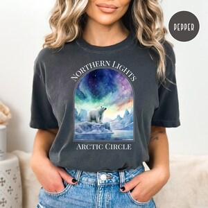 Op de afbeelding: Donkergrijs T-shirt met een aquarel illustratie van een ijsbeer op een ijsberg onder het Noorderlicht. De tekst "NORTHERN LIGHTS ARCTIC CIRCLE" staat boven en onder de afbeelding. Het shirt is Pepper van kleur.