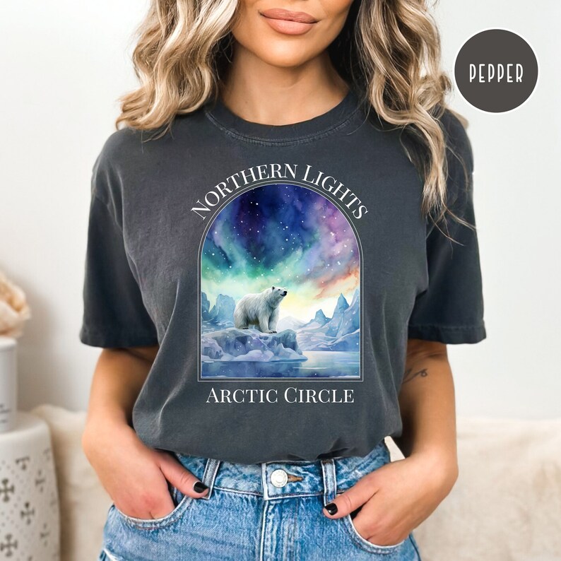 Op de afbeelding: Donkergrijs T-shirt met een aquarel illustratie van een ijsbeer op een ijsberg onder het Noorderlicht. De tekst "NORTHERN LIGHTS ARCTIC CIRCLE" staat boven en onder de afbeelding. Het shirt is Pepper van kleur.