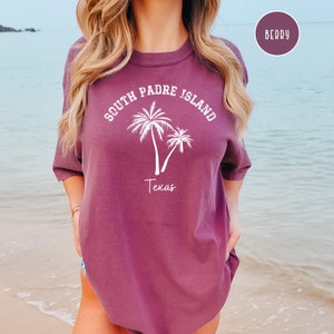 May include: A berry colored t-shirt with a white graphic design that says "South Padre Island Texas" with palm trees.