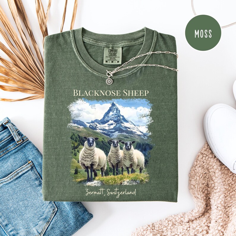 K&ouml;nnte beinhalten: Ein moosgr&uuml;nes T-Shirt mit einer Grafik von Schwarznasenschafen vor einer Bergkette. Der Text "BLACKNOSE SHEEP" und "Zermatt, Switzerland" sind auf das Shirt gedruckt. Eine silberne Halskette ist &uuml;ber das Shirt drapiert.