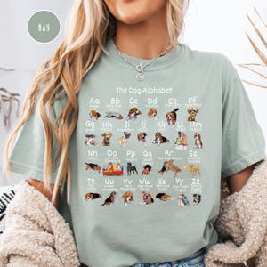 May include: A light green t-shirt with a graphic of a dog alphabet. Each letter of the alphabet is paired with a dog breed and a word describing the breed. The text "The Dog Alphabet" is at the top of the graphic.