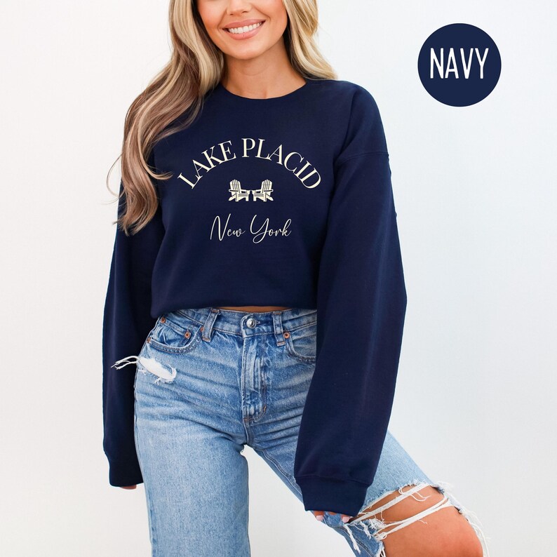 May include: Navy blue sweatshirt with the text "Lake Placid New York" in white script. The design includes two small white Adirondack chairs. A navy blue circle with the word "NAVY" is in the upper right corner.
