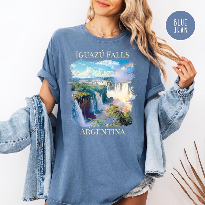 Pu&ograve; includere: T-shirt blu con un'immagine panoramica delle cascate dell'Iguaz&uacute; in Argentina. La grafica include il testo "Iguazu Falls" sopra l'immagine e "Argentina" sotto. La maglietta &egrave; di colore blu unito.