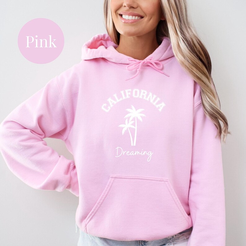 Puede incluir: Una sudadera con capucha rosa con un gr&aacute;fico blanco que dice "CALIFORNIA DREAMING" con palmeras.