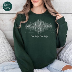 K&ouml;nnte beinhalten: Ein dunkelgr&uuml;nes Sweatshirt mit einer wei&szlig;en Grafik der Skyline von New York City, die im Wasser reflektiert wird. Der Text "New York, New York" ist unter der Grafik aufgedruckt.