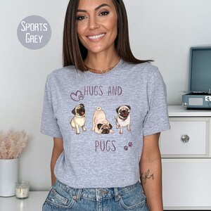 Puede incluir: Una camiseta gris con un gr&aacute;fico de tres carlinos y el texto "Hugs and Pugs" en morado.