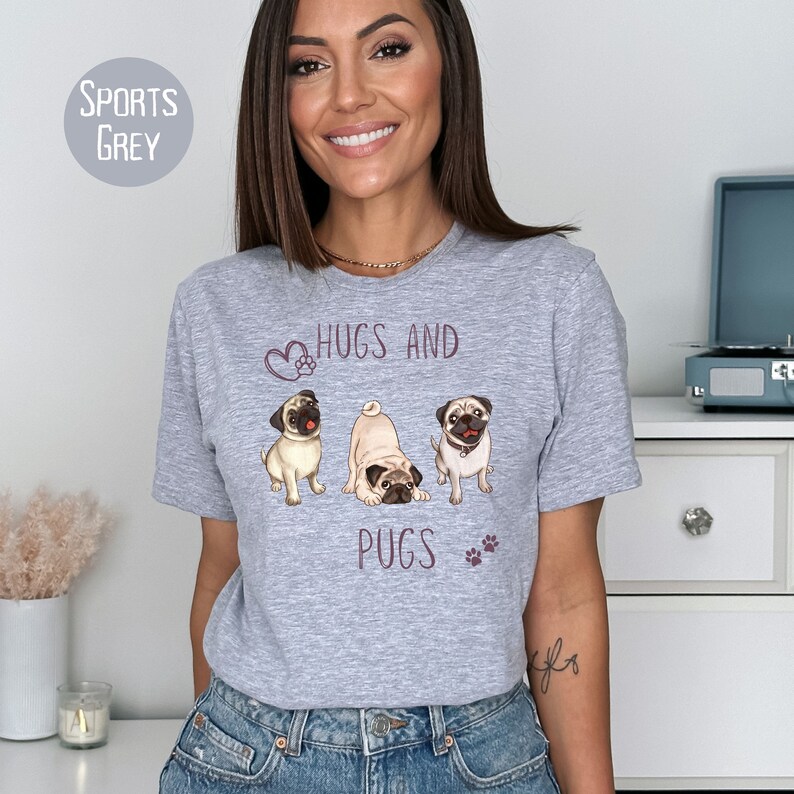Puede incluir: Una camiseta gris con un gr&aacute;fico de tres carlinos y el texto "Hugs and Pugs" en morado.