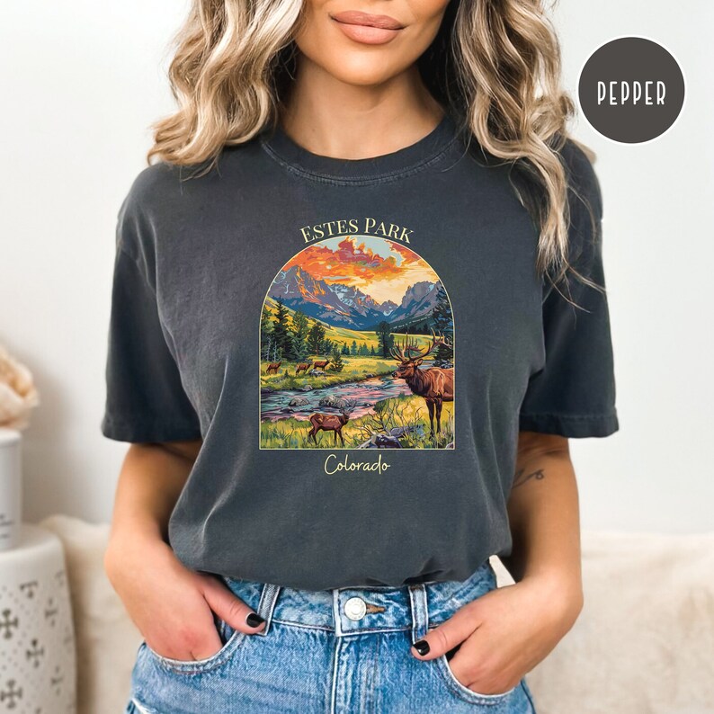May include: A dark grey t-shirt featuring a vintage-style illustration of a mountain landscape with a river and elk. The text "Estes Park Colorado" is printed on the shirt.