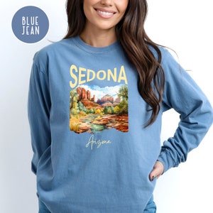 May include: A blue jean long-sleeve shirt with a graphic print of a landscape scene with red rock formations and a river. The text "SEDONA Arizona" is printed on the shirt.