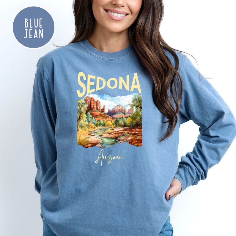 May include: A blue jean long-sleeve shirt with a graphic print of a landscape scene with red rock formations and a river. The text "SEDONA Arizona" is printed on the shirt.