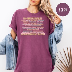 Pode incluir: Camiseta cor de berry com o texto "PRO-AMERICAN VALUES" e uma lista de princ&iacute;pios relacionados em fonte marrom claro. A pessoa que usa a camiseta tem cabelo loiro e uma jaqueta jeans.