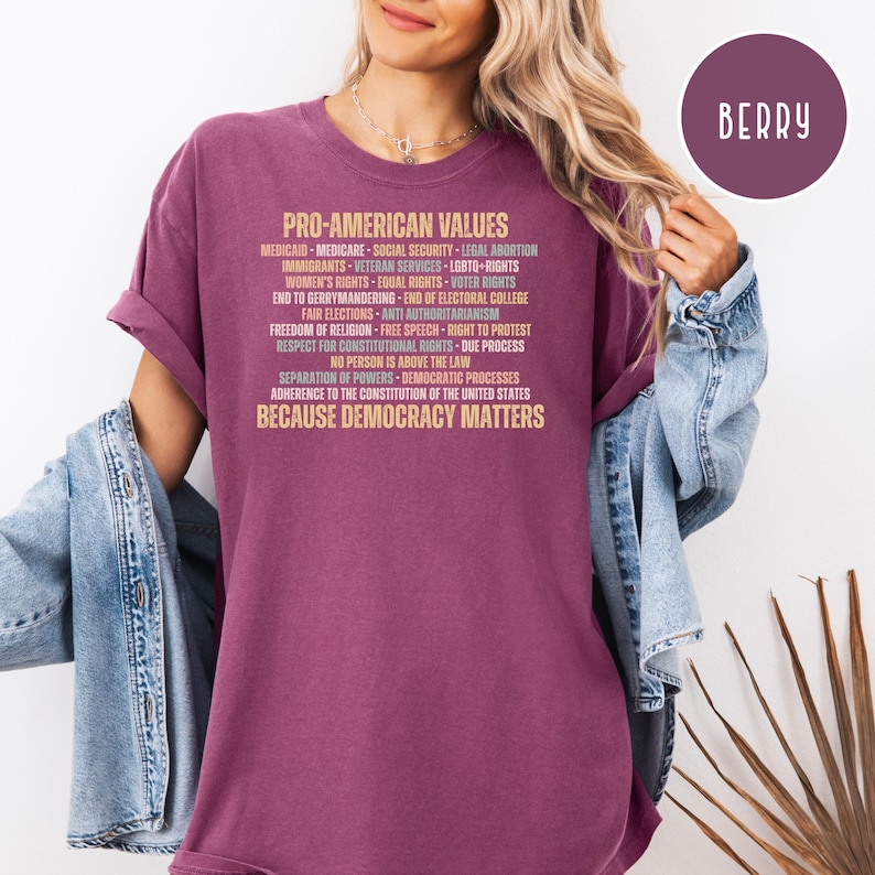 Pode incluir: Camiseta cor de berry com o texto "PRO-AMERICAN VALUES" e uma lista de princ&iacute;pios relacionados em fonte marrom claro. A pessoa que usa a camiseta tem cabelo loiro e uma jaqueta jeans.