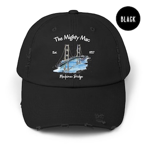 Może przedstawiać: Czarna, zniszczona czapka z daszkiem z białym nadrukiem mostu Mackinac, z tekstem "The Mighty Mac" i "Est. 1957 Mackinac Bridge".