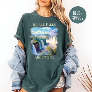 Pu&ograve; includere: Una maglietta verde scuro con un'immagine panoramica delle cascate dell'Iguaz&uacute; in Argentina. La grafica include il testo "IGUAZ&Uacute; FALLS" sopra le cascate e "ARGENTINA" sotto. La maglietta ha una vestibilit&agrave; rilassata.
