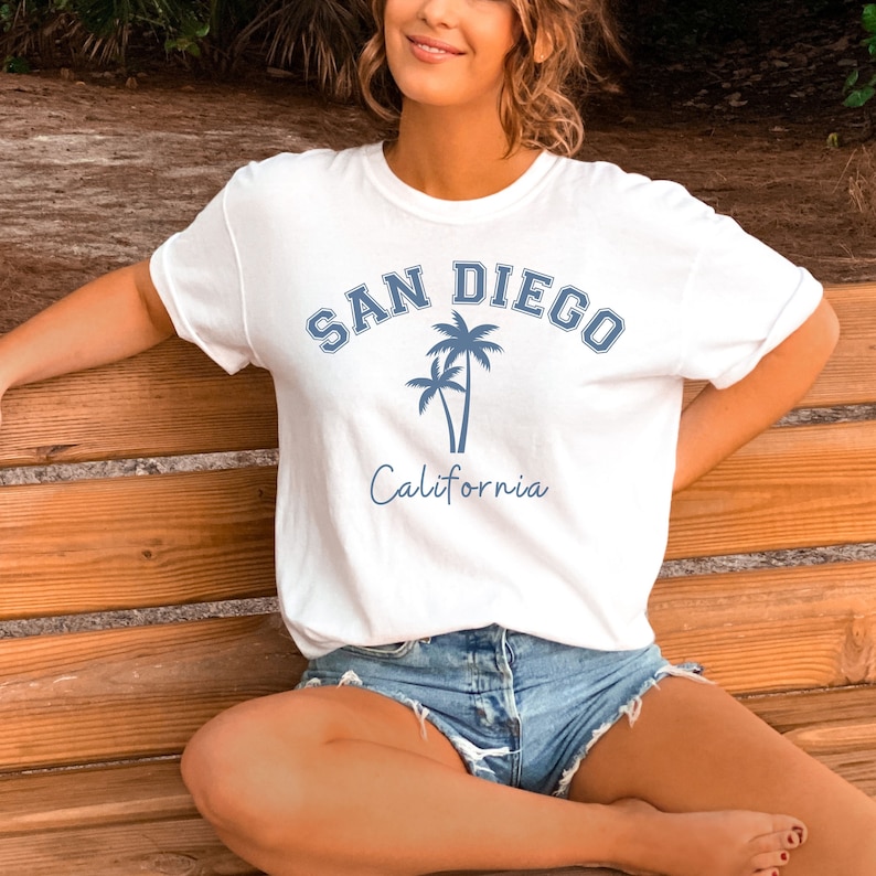 Może przedstawiać: Biała koszulka z niebieskim nadrukiem palm i tekstem "SAN DIEGO California".