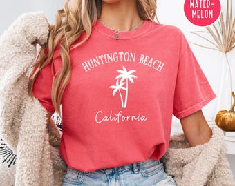 May include: A red t-shirt with the text "Huntington Beach California" and a palm tree graphic. The text is white. The shirt is worn by a person with long blonde hair.
