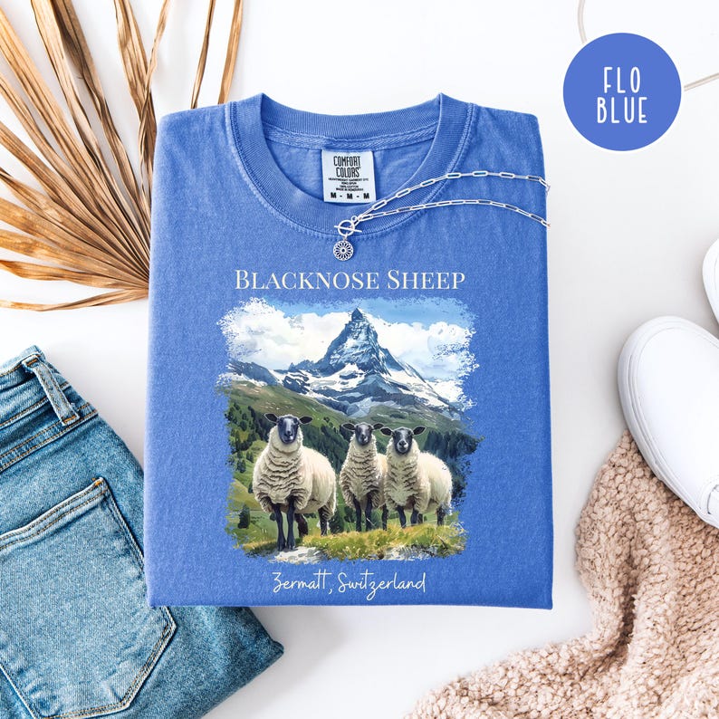K&ouml;nnte beinhalten: Ein blaues T-Shirt mit einer Grafik von drei Schwarznasenschafen vor einer Berglandschaft. Der Text "BLACKNOSE SHEEP" und "Zermatt, Switzerland" sind auf dem Shirt aufgedruckt. Eine silberne Halskette liegt auf dem Shirt.