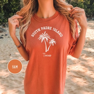 May include: A burnt orange t-shirt with white text that reads "South Padre Island Texas" and a graphic of two palm trees.
