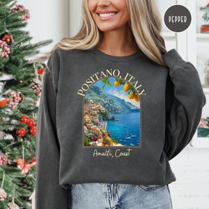 K&ouml;nnte beinhalten: Ein anthrazitfarbener Sweatshirt mit einem grafischen Druck einer farbenfrohen Szene von Positano, Italien, mit dem Text "Positano, Italien" und "Amalfik&uuml;ste" in gelber Schrift.