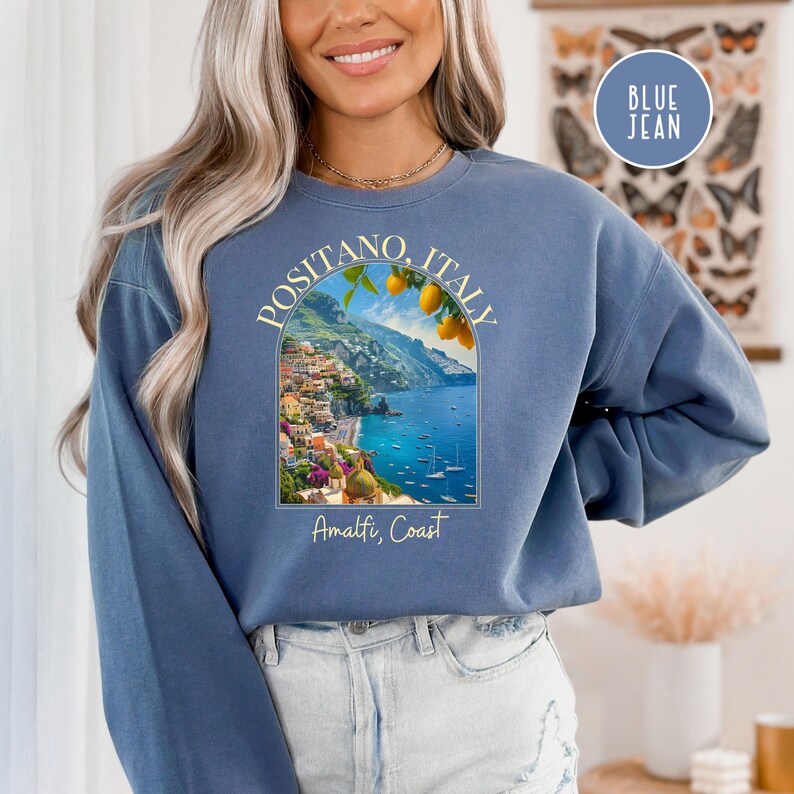 K&ouml;nnte beinhalten: Ein blaues Sweatshirt mit einem Aufdruck von Positano, Italien, mit einer farbenfrohen Stadtlandschaft, einem blauen Meer und einem Zitronenbaum. Der Text "Positano, Italien" und "Amalfik&uuml;ste" ist auf dem Sweatshirt aufgedruckt.