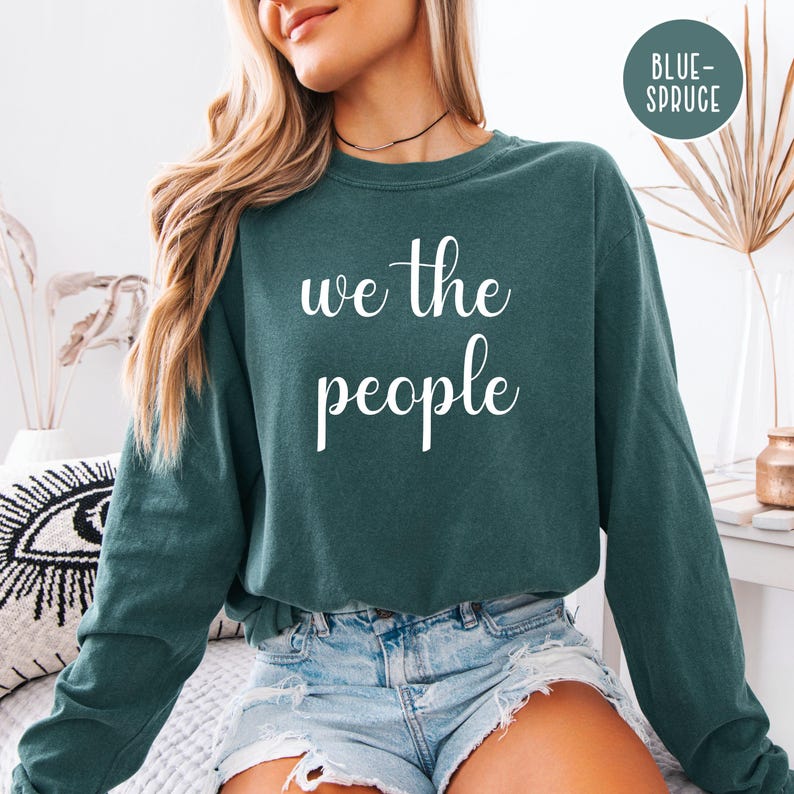 Puede incluir: Una camiseta de manga larga azul turquesa con el texto "we the people" en letras blancas.