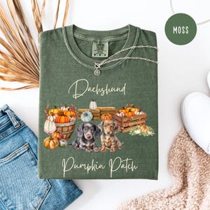May include: Moss green Comfort Colors t-shirt with a Dachshund Pumpkin Patch design. The graphic features two dachshunds, pumpkins, and the text "Dachshund Pumpkin Patch". A silver necklace with a pendant is draped on the shirt.