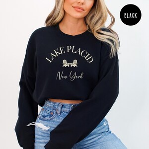 May include: Black sweatshirt with the text "Lake Placid New York" in a cream-colored font. The design includes two small chair illustrations. A black circle with the word "BLACK" is in the upper right corner.