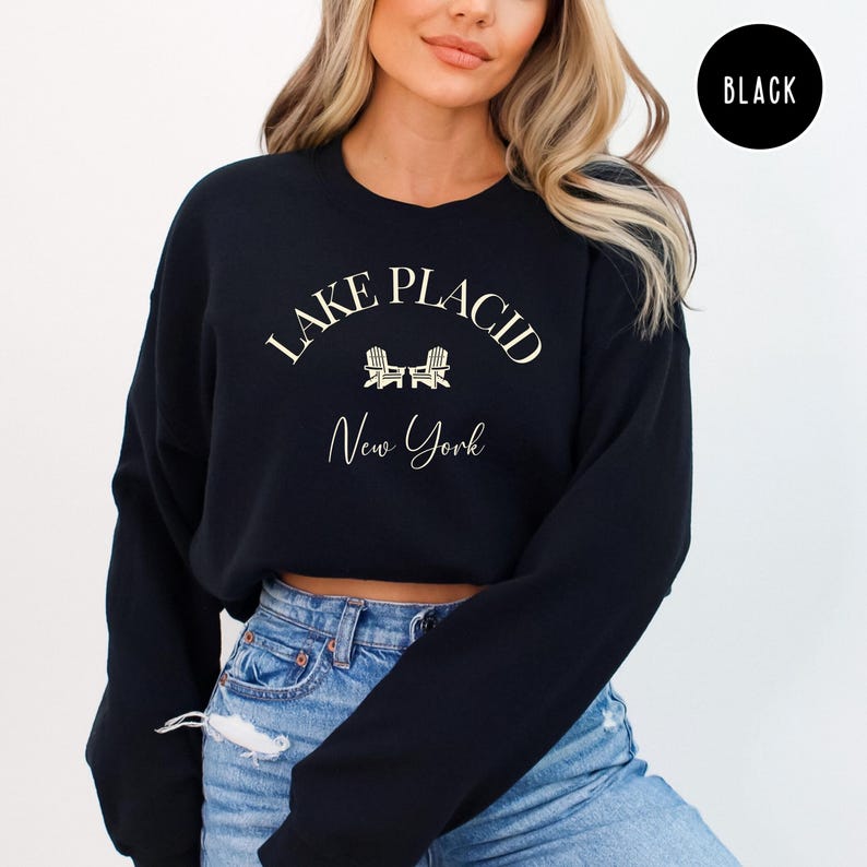 May include: Black sweatshirt with the text "Lake Placid New York" in a cream-colored font. The design includes two small chair illustrations. A black circle with the word "BLACK" is in the upper right corner.
