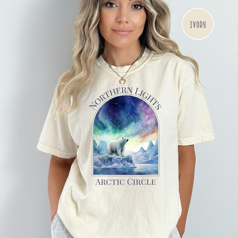 Op de afbeelding: Ivoor kleurig T-shirt met een aquarel ontwerp van een ijsbeer op een ijsberg onder het noorderlicht. Het ontwerp bevat de tekst "NORTHERN LIGHTS" erboven en "ARCTIC CIRCLE" onder de afbeelding.
