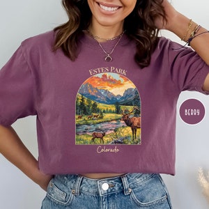 May include: A purple t-shirt featuring a graphic of a mountain scene with a sunset and a deer. The text "Estes Park Colorado" is printed on the shirt.