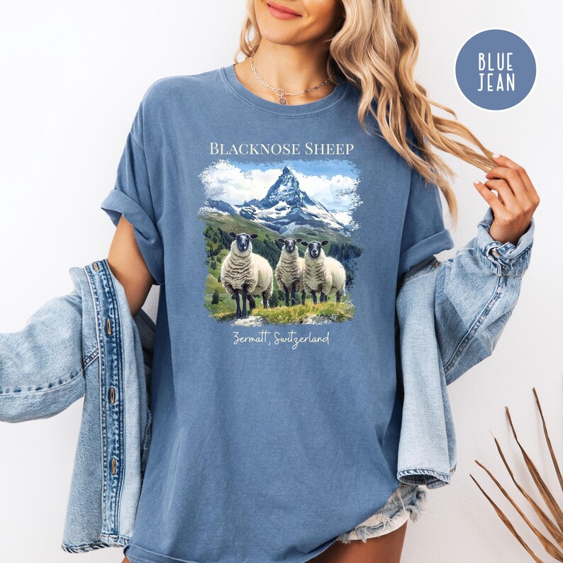 K&ouml;nnte beinhalten: Blaues T-Shirt mit einer Grafik von drei Schwarznasenschafen vor dem Matterhorn in Zermatt, Schweiz. Der Text "BLACKNOSE SHEEP" und "Zermatt, Switzerland" sind oberhalb und unterhalb des Bildes aufgedruckt.