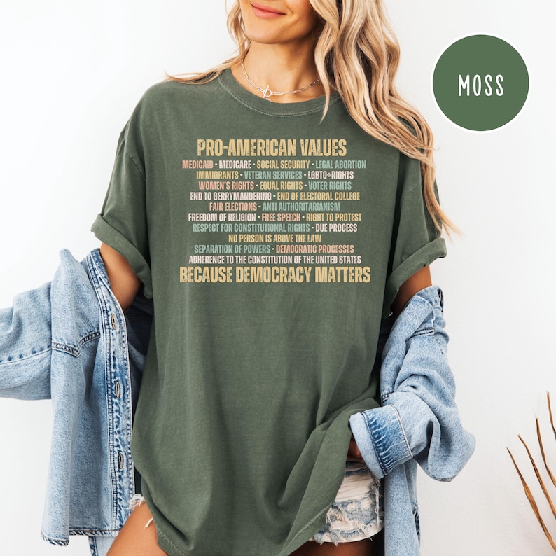 Pode incluir: Uma camiseta verde-oliva com o texto "PRO-AMERICAN VALUES" e uma lista de valores em uma cor contrastante. A camisa tamb&eacute;m tem o texto "BECAUSE DEMOCRACY MATTERS". Uma etiqueta circular "MOSS" est&aacute; no canto superior direito.