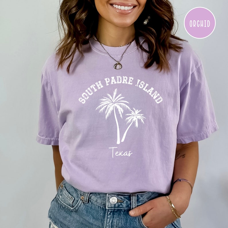 May include: A lavender colored t-shirt with the text "South Padre Island Texas" and two palm trees in white.
