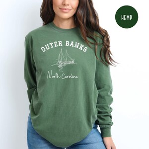 May include: Green long-sleeve sweatshirt with the text "Outer Banks North Carolina" and a sailboat graphic. The word "Hemp" is printed on a green circle. The sweatshirt is made of a soft material.