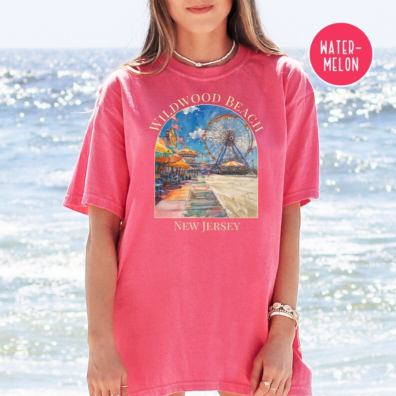 May include: A pink t-shirt with a graphic of a boardwalk scene in Wildwood, New Jersey. The graphic includes a Ferris wheel, beach umbrellas, and the text "Wildwood Beach, New Jersey".