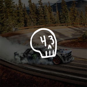 May include: A grey rally car drifting on a mountain road, emitting thick white tyre smoke. A white hand-drawn skull graphic with the number 43 is centred over the car. The vehicle features visible sponsor logos and dark wheels.