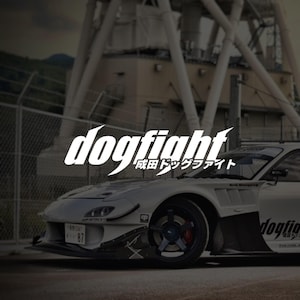 May include: A white sports car with a modified front bumper, vented fenders, and dark multi-spoke wheels. The text "dogfight" and "成田ドッグファイト" are displayed in white in the centre of the image over the car and a blurred industrial background.