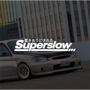 May include: A beige saloon car with a dark bonnet and gold-rimmed wheels, featuring a white decal on the windscreen that reads "Superslow" in English and Japanese characters, with a California number plate displaying the text "ATS VEE" on the front bumper.