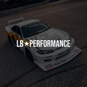 Peut inclure: Une voiture de course blanche LB PERFORMANCE dotée d'un kit carrosserie large, d'un grand aileron arrière et de divers autocollants de sponsors dont LIBERTY WALK, LIQUI MOLY et HUG HUG. Le véhicule affiche le numéro 23 et des roues noires.