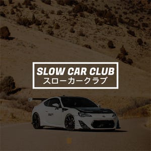 Könnte beinhalten: Ein weißer Sportwagen mit schwarzer Motorhaube und Heckflügel auf einer Straße. Ein weißes rechteckiges Schild mit schwarzem Rand zeigt den Text SLOW CAR CLUB und japanische Schriftzeichen in Weiß, zentriert über dem Auto vor einer Hügellandschaft.