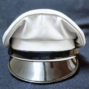 以下が含まれることがあります： 質感のある表面を持つ白いキャップで、前面には黒いパテントレザーのバンドと、お揃いの黒いパテントレザーのバイザーが付いています。キャップは構造化されたクラウンと丸みを帯びたトップを備えており、すっきりとしたコントラストのある外観を呈しています。