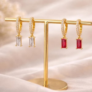 May include: Gold-toned hoop earrings with clear and red rectangular pendants. The hoops are encrusted with small, sparkling stones. The earrings are displayed on a gold jewelry stand against a soft, neutral background.