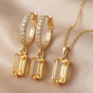 Puede incluir: Un conjunto de joyas doradas con un par de pendientes de aro y un collar con colgante. Los pendientes tienen una fila de pequeñas piedras transparentes a lo largo del aro y una gema amarilla rectangular. El collar tiene un colgante de gema amarilla a juego.