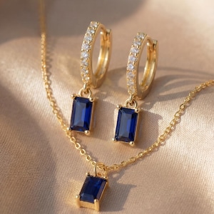 Könnte beinhalten: Ein goldfarbenes Schmuckset mit Halskette und Ohrringen. Die Ohrringe sind im Creolen-Stil gehalten, mit kleinen, klaren Steinen besetzt und haben einen rechteckigen, tiefblauen Edelstein. Die Halskette hat einen passenden blauen Edelsteinanhänger.