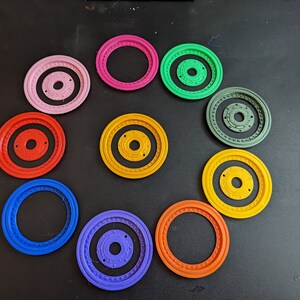 May include: Ten colorful plastic rings with different designs and colors, including red, blue, green, yellow, pink, and purple. The rings are arranged in a circle on a black surface.
