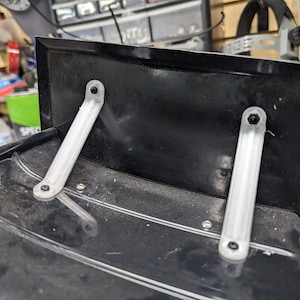 May include: Two silver metal brackets are attached to a black plastic surface with screws. The brackets are angled and have a slight curve to them.