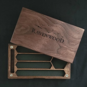 May include: A wooden box with a hinged lid, featuring the engraved text "Ravenwood" and a flock of birds. The box is open, revealing a green felt interior with several hexagonal compartments. The box is made of dark wood.